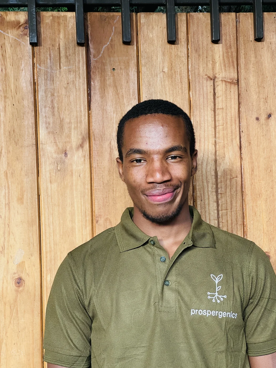 Finehas Mpoe smiling, wearing an olive Prospergenics polo shirt in front of a wooden fence